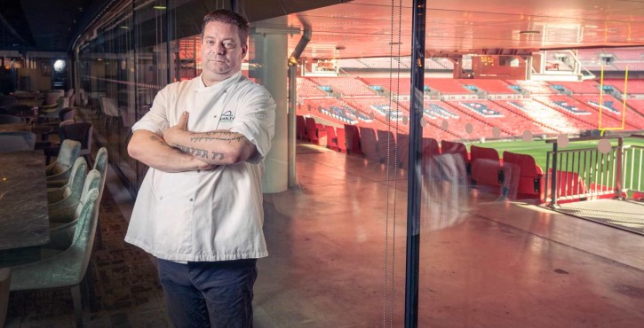 Chef Stefan Pappert standing with crossed arms in a luxury dining area overlooking a stadium with empty red seats, reflecting through the glass wall.