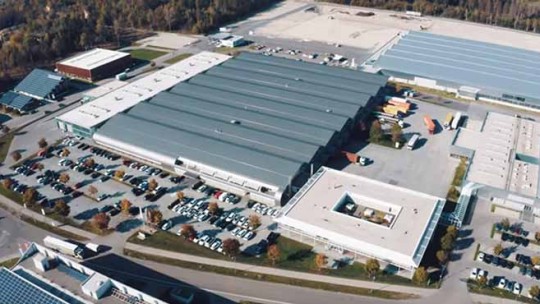 Et fugleperspektiv av en stor industribygning med en parkeringsplass og flere kjøretøy.