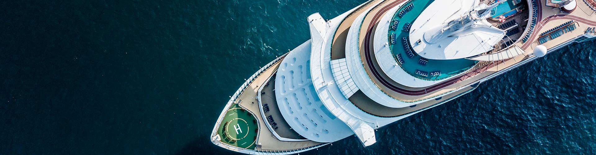An aerial view of a large cruise ship sailing on blue ocean waters, featuring a helipad on the deck.