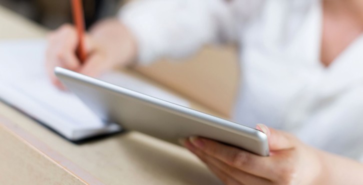 Personne tenant une tablette et prenant des notes au crayon sur un carnet dans un contexte d’apprentissage ou de travail.