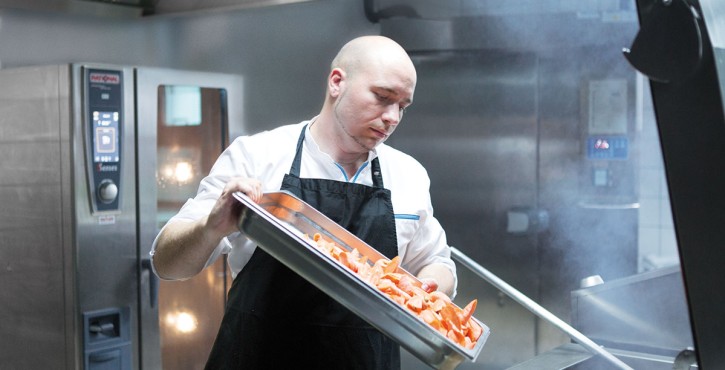 A chef in a professional kitchen carefully handling a tray of chopped vegetables with steam rising in the background.