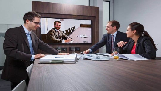 Three business professionals in a meeting room having a video conference with a fourth person displayed on a screen.