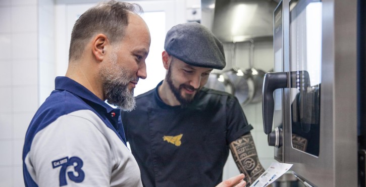 Two men in a kitchen discussing the features of a RATIONAL iCombi Pro oven, one holding a paper with details.