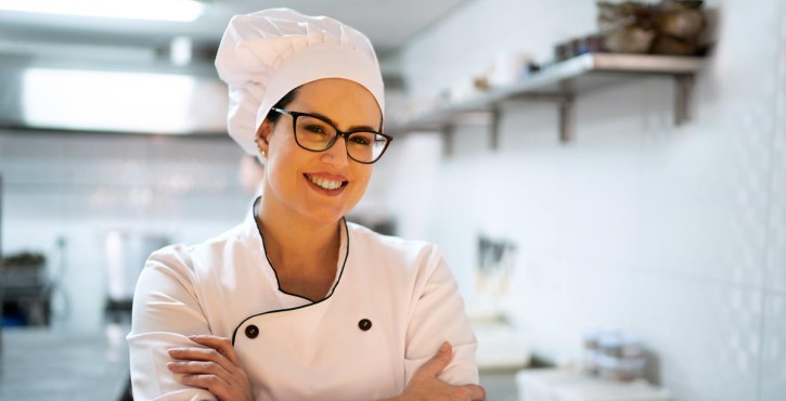 Une cheffe sourit, les bras croisés dans une cuisine, portant une veste de chef blanche et un toque.