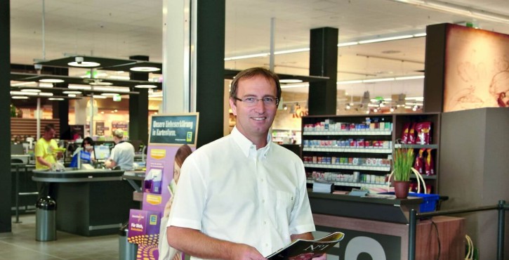 Un homme se tient dans un couloir de magasin tenant une brochure, avec des étagères et des clients en arrière-plan.