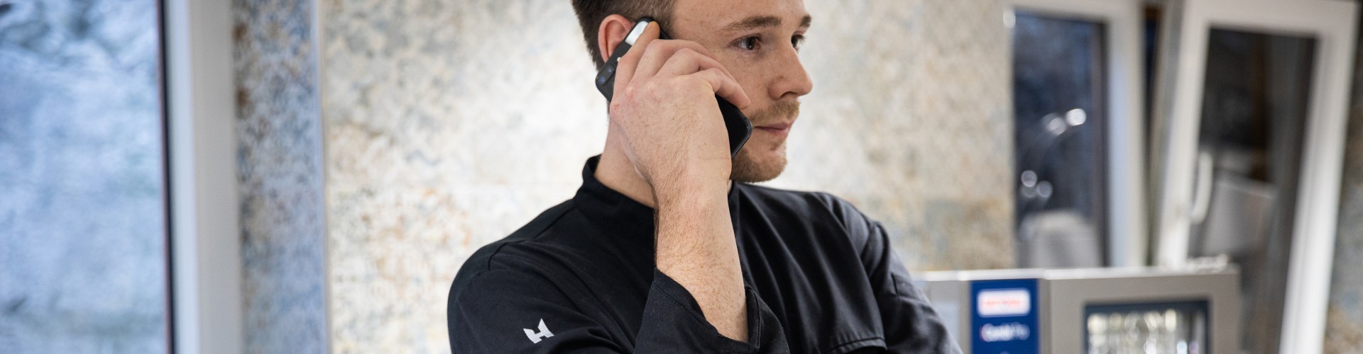 Chef con uniforme negro hablando por teléfono en una cocina profesional.