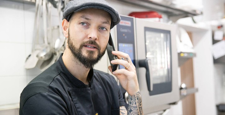 Bearded man with tattoos talking on the phone in a kitchen beside a combi-steamer.