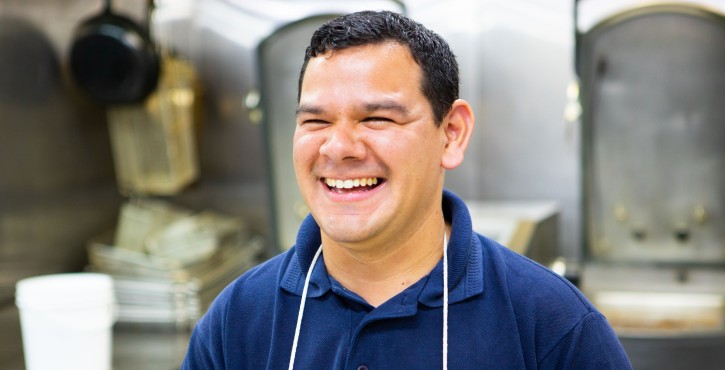 Smiling chef in a blue polo shirt and white apron in a professional kitchen.