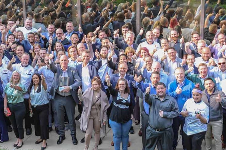 Foto grupal grande de empleados de Rational USA en ropa de negocios y uniformes de chef, al aire libre, levantando el pulgar frente a un edificio de vidrio.