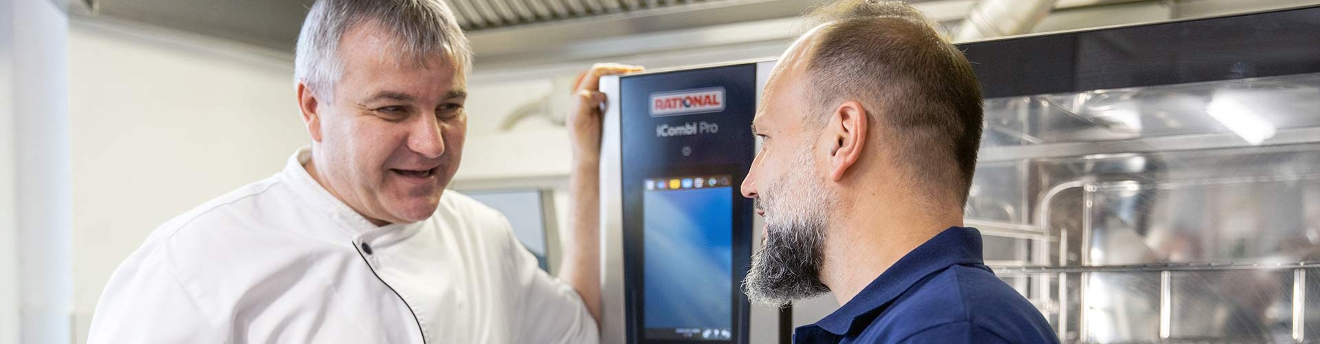 Two men in a kitchen discussing in front of a RATIONAL iCombi Pro, one wearing a chef‘s jacket and the other in a blue shirt.