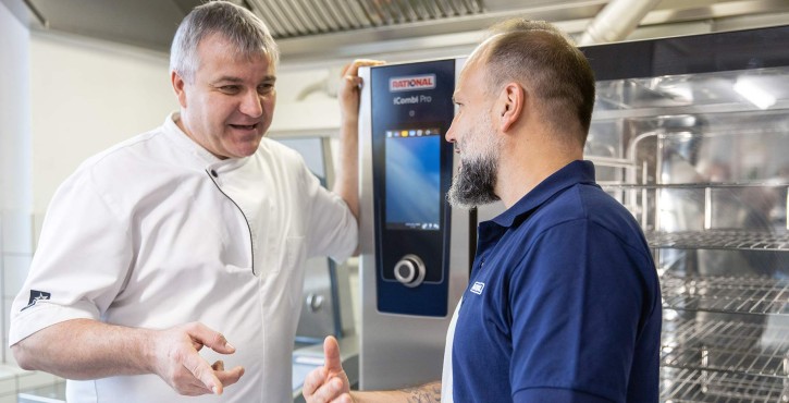 Two men in a kitchen discussing in front of a RATIONAL iCombi Pro, one wearing a chef's jacket and the other in a blue shirt.