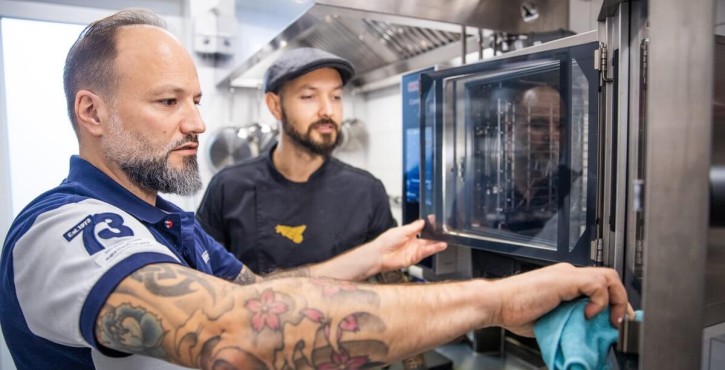 Two men in a kitchen, one explains how to use a RATIONAL oven, while the other adjusts it.