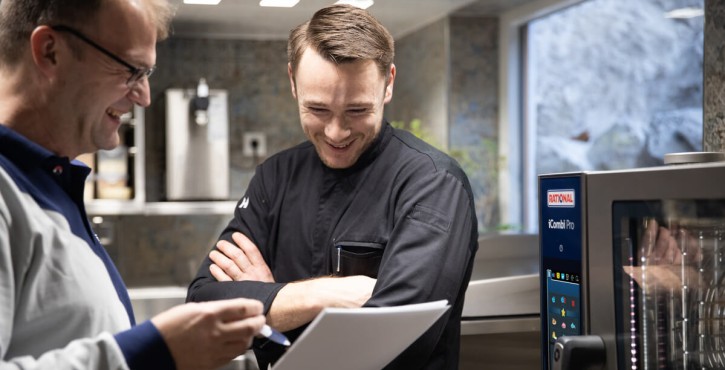 Two men check a document during a technical service meeting in front of a Rational iCombi Pro oven.