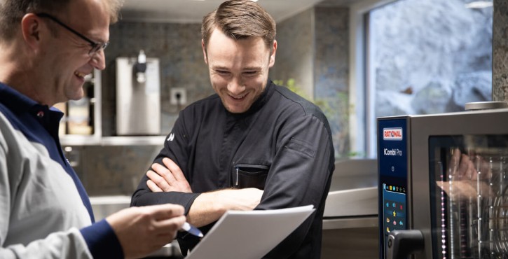 Two men smiling and discussing a document in a kitchen beside an iCombi Pro combi-steamer.