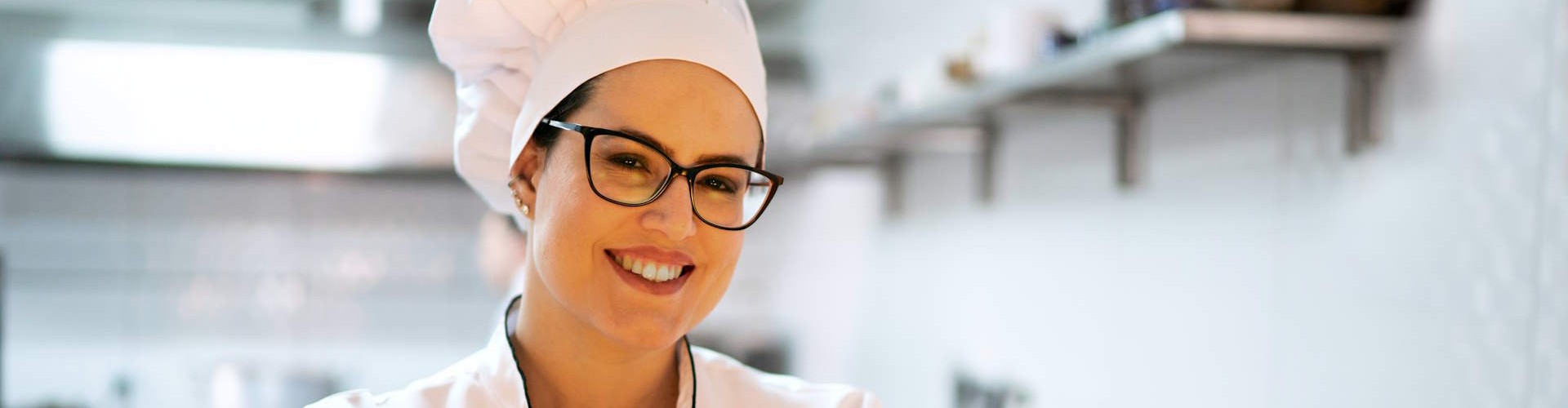 Eine Köchin mit Brille und weißer Kochmütze lächelt selbstbewusst, während sie mit verschränkten Armen in einer Küche steht.