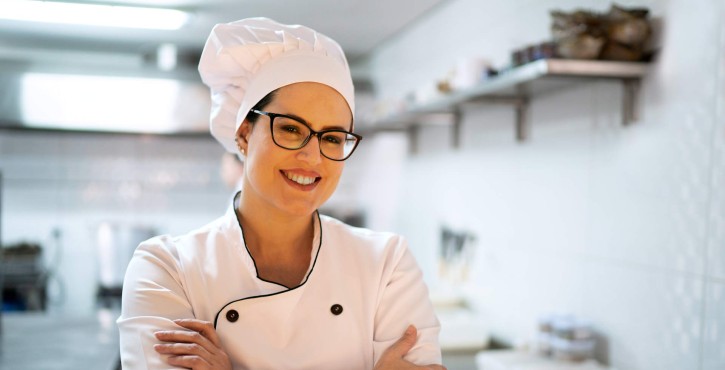 A female chef with glasses and a white chef's hat smiles confidently while standing with crossed arms in a kitchen.