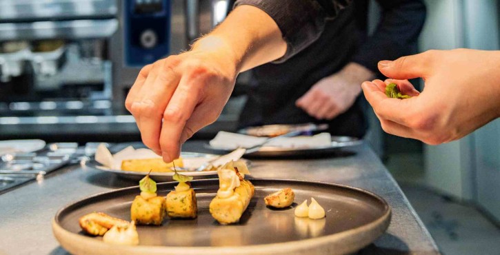 A chef in a black uniform decorates a plate in the kitchen, adding finishing touches to gourmet dishes under pendant lights.