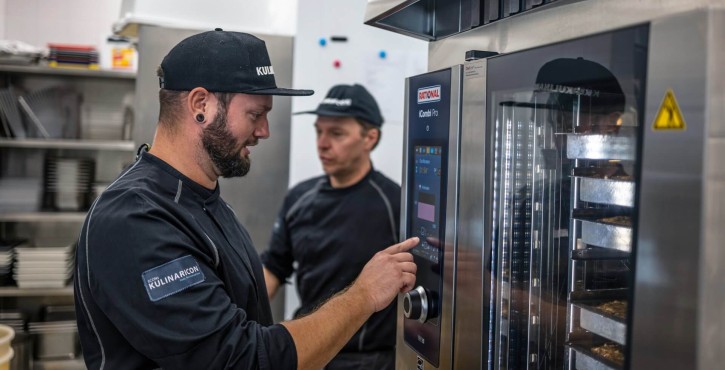 Two chefs in a kitchen, one adjusting the settings on a Rational iCombi Pro oven while the other looks on.