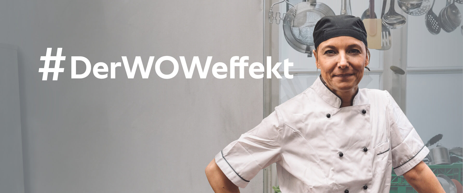 Eine weibliche Köchin in weißer Uniform und schwarzem Kochhut steht selbstbewusst mit verschränkten Armen neben dem Text '#TheWOWeffect' in einer mit Kochutensilien gefüllten Küche.