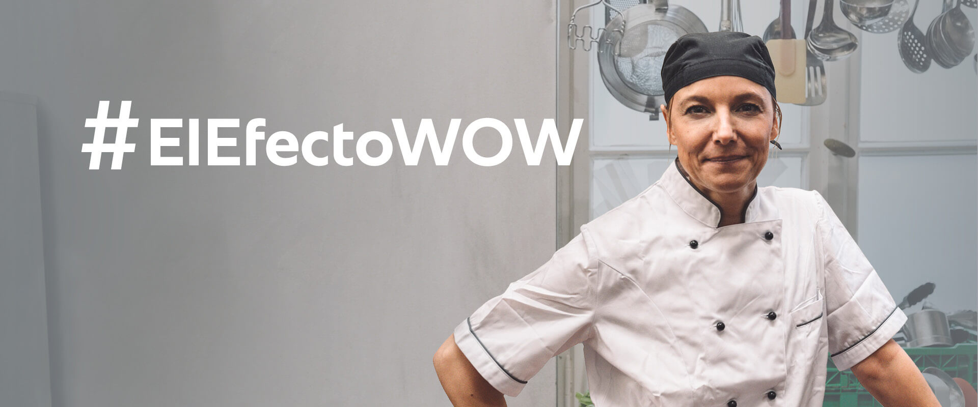 Una chef femenina con uniforme blanco y un gorro de chef negro se muestra confiada con los brazos cruzados, junto al texto '#TheWOWeffect' en una cocina llena de utensilios de cocina.