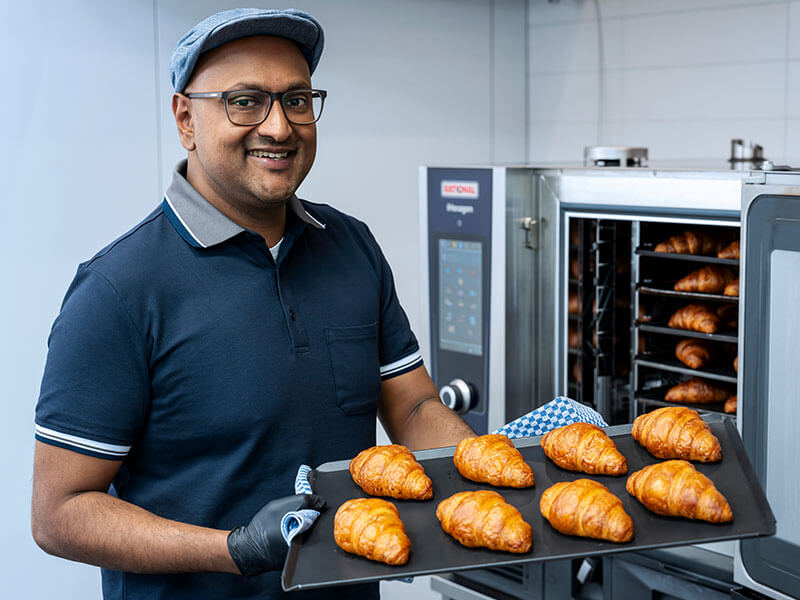 Lächelnder Mann hält ein Blech mit frisch gebackenen Croissants vor einem RATIONAL iHexagon Kombidämpfer.