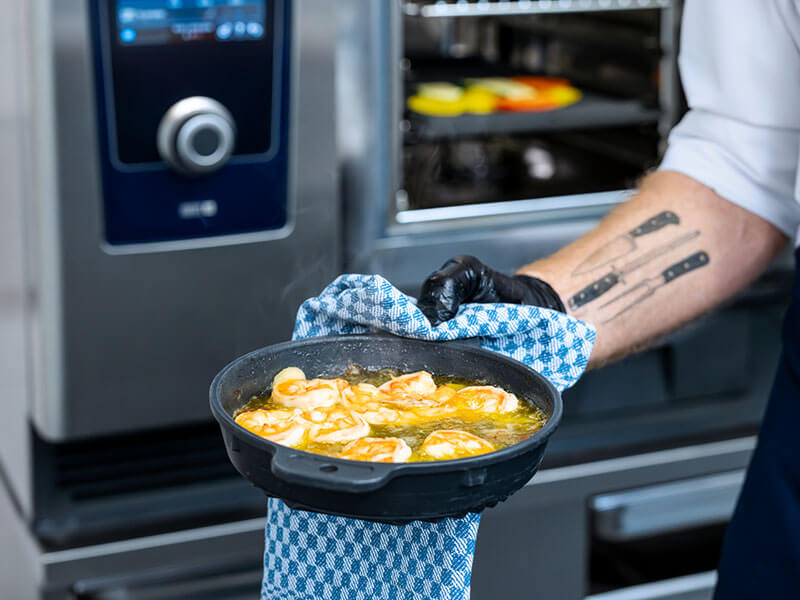 Koch hält eine heiße Pfanne mit brutzelnden Garnelen vor einem RATIONAL iHexagon Kombidämpfer.