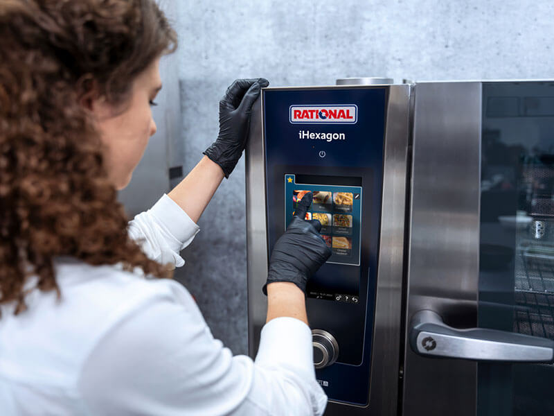 Köchin mit schwarzen Handschuhen wählt ein Kochprogramm auf dem Touchscreen des RATIONAL iHexagon Kombidämpfers.