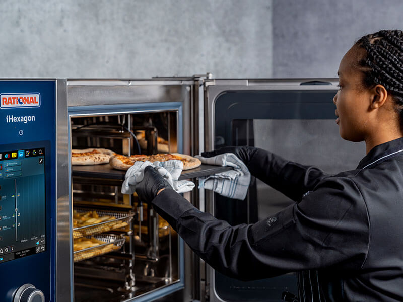 Köchin in schwarzer Uniform entnimmt frisch gebackene Pizzen aus einem RATIONAL iHexagon Kombidämpfer.