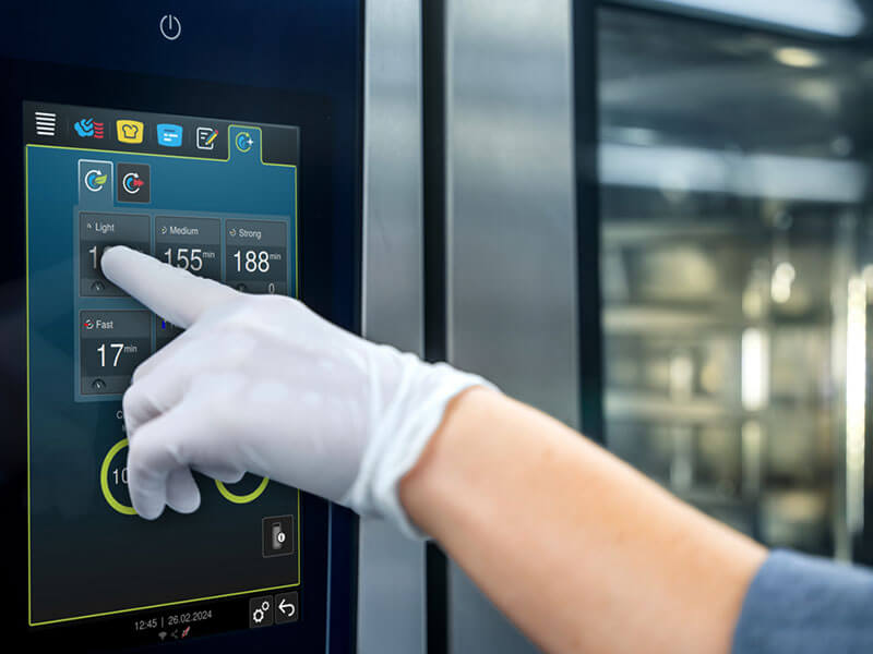Behandschuhte Hand wählt ein Reinigungsprogramm auf dem Touchscreen des RATIONAL iHexagon Kombidämpfers.