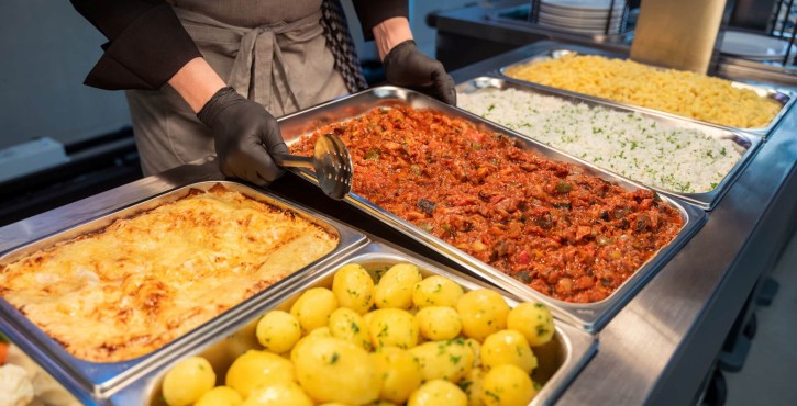Buffet with trays of food including potatoes, lasagna, meat stew, rice, and pasta. Staff member serving with tongs.