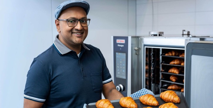 Smiling man holding a tray of freshly baked croissants in front of a RATIONAL iHexagon combi oven.