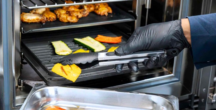 Koch in schwarzen Handschuhen entnimmt gegrilltes Gemüse aus einem RATIONAL Kombidämpfer mit einer Zange.