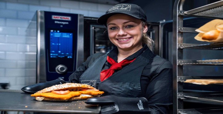 Smiling chef holding a tray with a crispy schnitzel sandwich in front of a RATIONAL iHexagon combi oven.