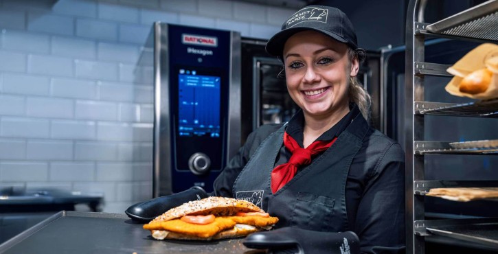 
Smiling chef holding a breaded schnitzel sandwich in front of a RATIONAL iHexagon combi oven.