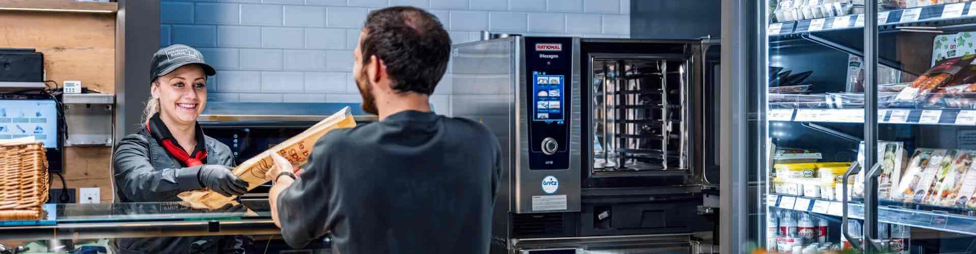 Eine Kassiererin reicht einem Kunden eine Tüte mit Lebensmitteln vor einem RATIONAL Kombidämpfer, im Hintergrund eine Vitrine mit Produkten.