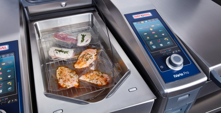 A Rational iVario Pro 2-S with various vacuum-sealed meat items inside, displayed on a stainless steel countertop.