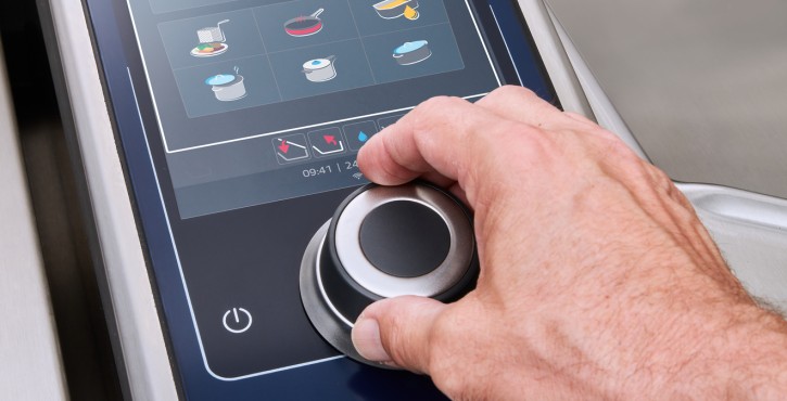 A close-up of the Rational iVario Pro cooking appliance's touchscreen panel, showing a hand interacting with the rotary control.