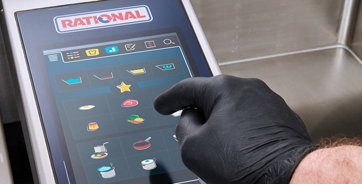 Un primer plano de una mano con guante negro interactuando con el panel de control táctil de un aparato Rational iVario Pro.