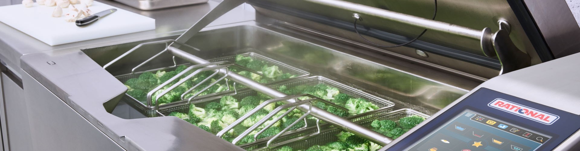 A professional kitchen showing an open cooking unit with broccoli being prepared in baskets, featuring a digital control panel.