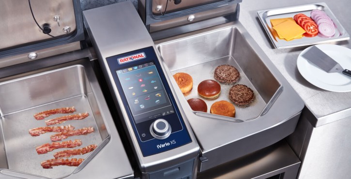 The iVario Pro advanced bratt pan from RATIONAL has two cooking pans, one side is cooking crispy bacon the other side is cooking burgers and buns.
