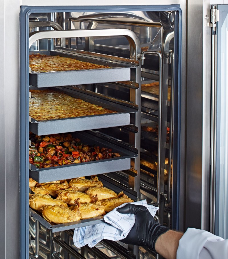A chef in a white uniform and black gloves is pulling out a tray of chicken from a Rational iCombi Pro oven, with additional trays of vegetables and pasta visible inside.