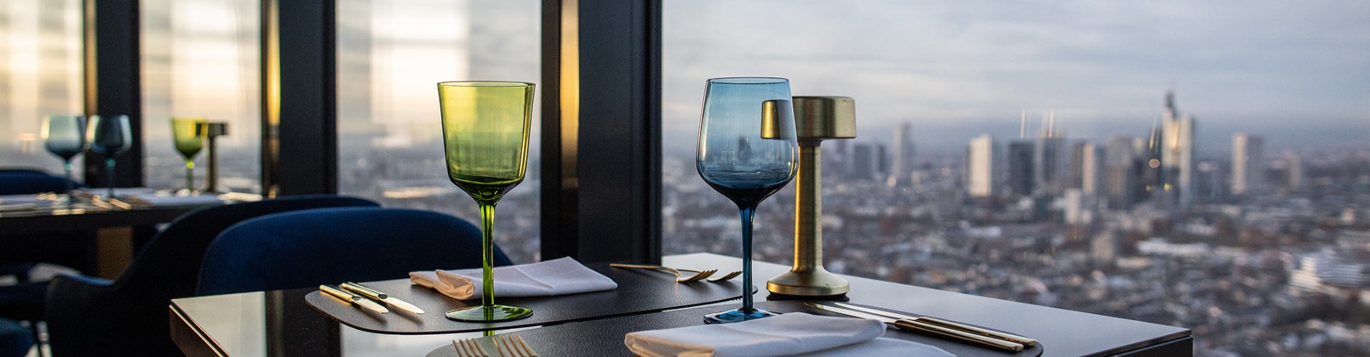 Mesa de restaurante elegante con copas verdes y azules, cubiertos y un portavelas, con vista al horizonte de la ciudad.