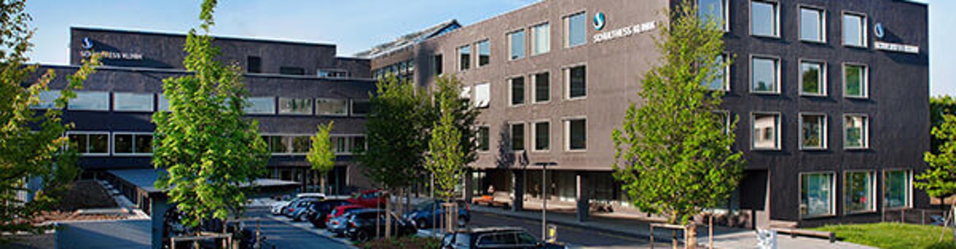 The exterior of Schulthess Klinik, a modern medical facility with greenery, parking, and a clear blue sky.