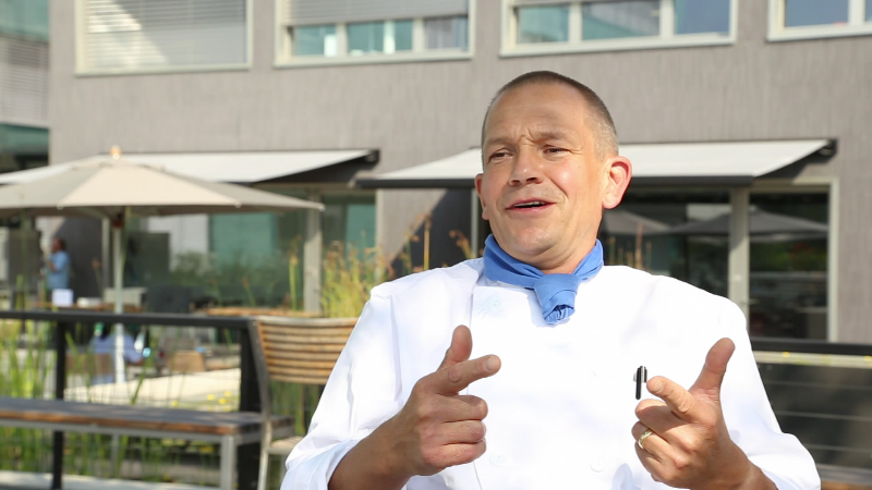 A chef in a white uniform and blue neckerchief, smiling and gesturing with his hands in front of a modern building with outdoor seating.