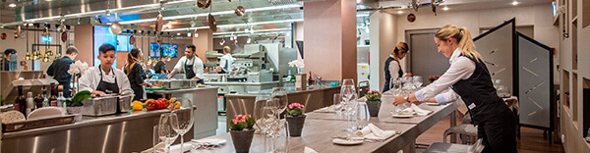 Waitress setting a table in a modern open kitchen restaurant with chefs preparing food.