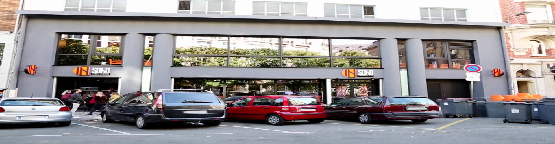 The front of a building with large windows, a logo, and cars parked outside.