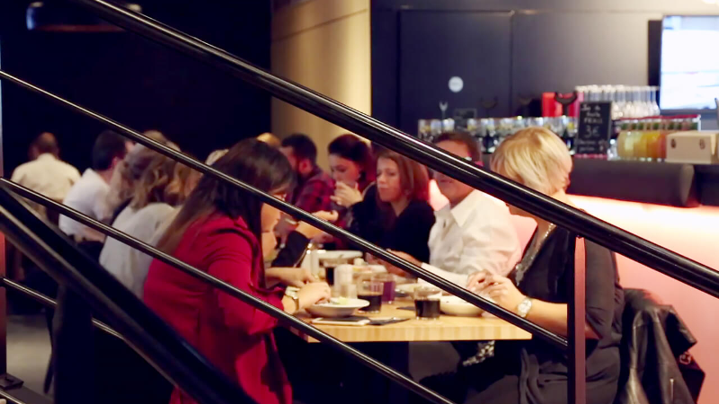 People dining at a restaurant with a focus on a woman in a red jacket and a person in a black shirt.