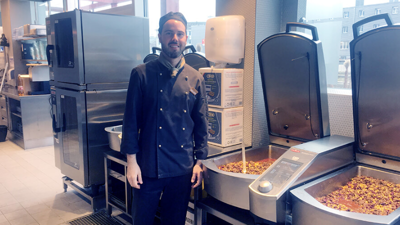 A chef in a black uniform stands next toa VarioCookingCenter with food, in a modern kitchen setup with Rational ovens in the background.