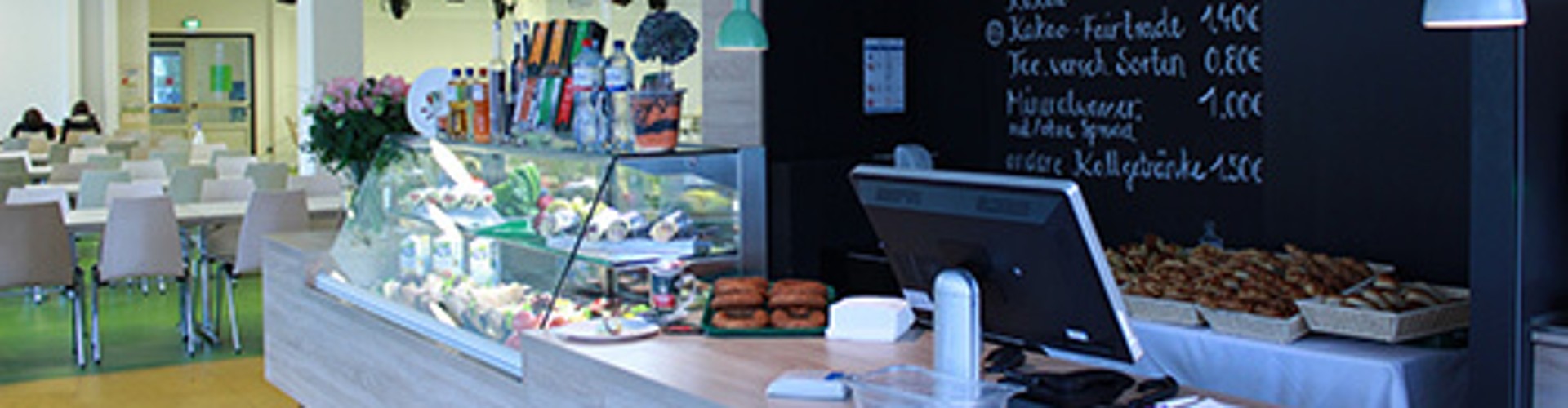 Interior of Café Lounge featuring a counter with a display of pastries and cookies, a chalkboard menu on the wall, and several tables in the background. Bright, inviting atmosphere.