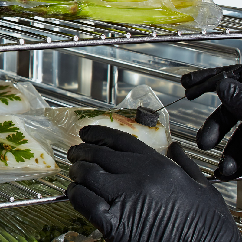 Gloved hand inserting a temperature probe into a vacuum-sealed food pouch on a wire rack.